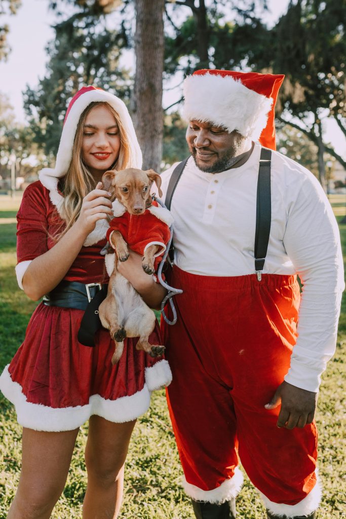 A joyful couple in Santa costumes holds a dog in a park, celebrating Christmas.